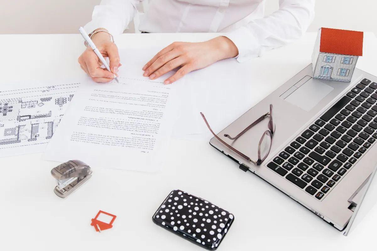 Eine Hand unterschreibt ein Dokument am Schreibtisch neben einem Laptop